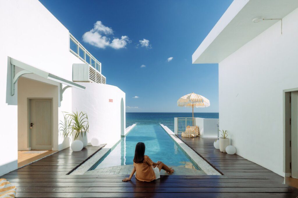A person sits by a narrow pool between white buildings, facing the sea. There are a few plants, a door, and an umbrella under a clear blue sky with small clouds.