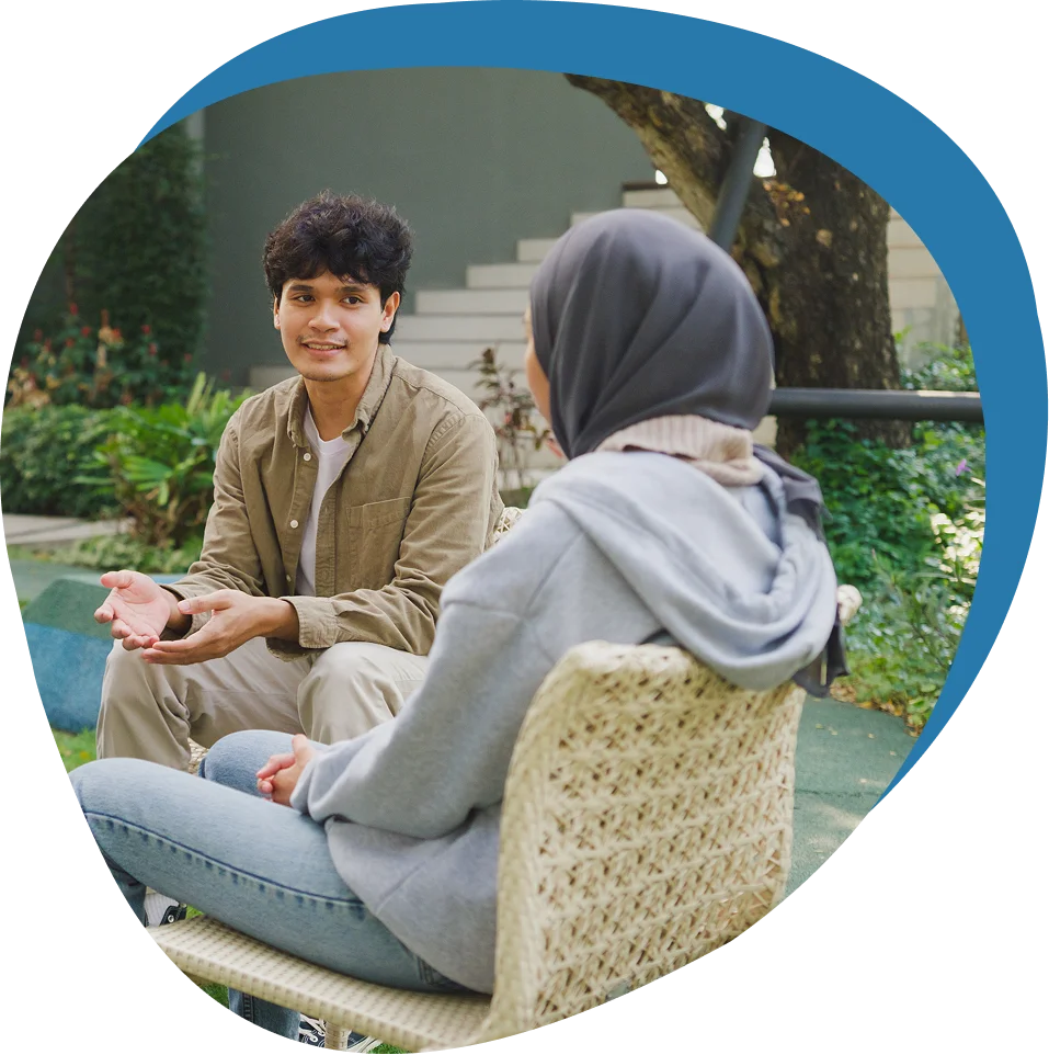 A man and a woman sit outdoors on wicker chairs, engaged in conversation, with greenery in the background.