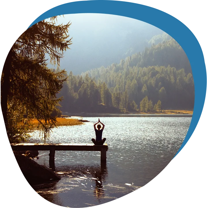 Person sitting cross-legged on a wooden dock by a lake, meditating with hands raised, surrounded by trees and mountains under a clear sky.